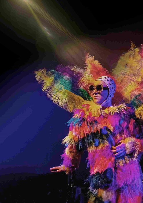 "Young Elton" dressed in a multi-coloured, feathered jacket and trousers with big round sunglasses. On the back of the jacket, surrounding his head, are large, brightly coloured feathers matching the jacket and trousers. 