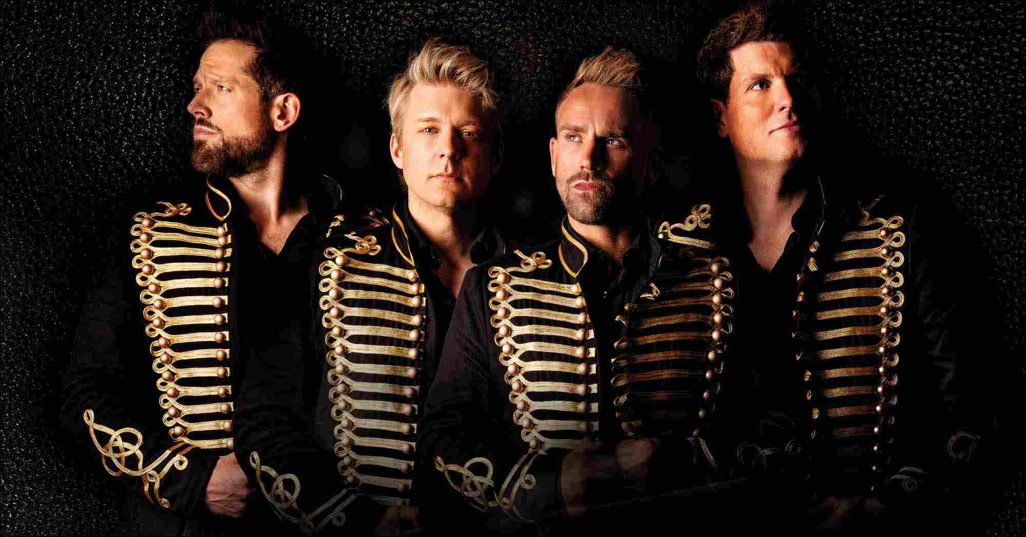 Four men wearing black jackets with ornate gold detailing stand against a dark background, looking in different directions with confident, dramatic expressions.