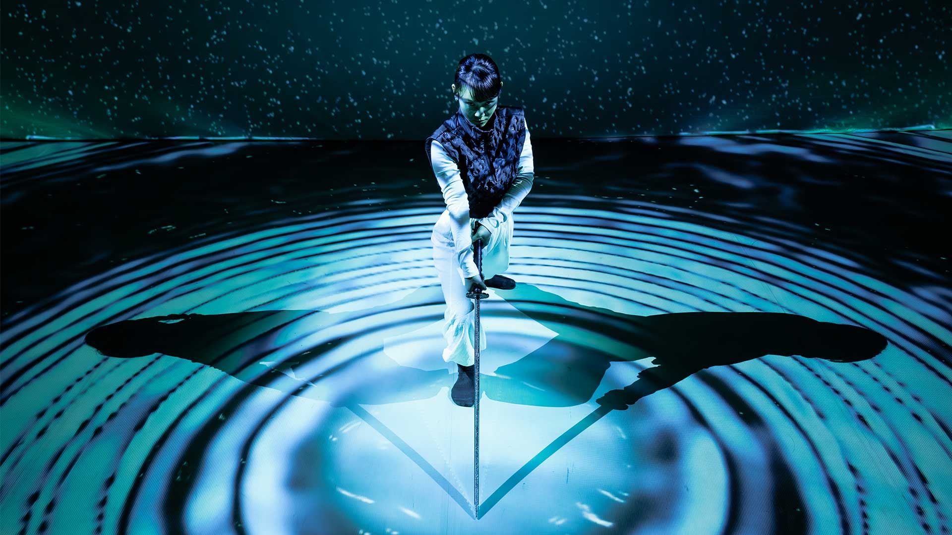 An image of a dancer with a sword amid blue circular light patterns and shadows.