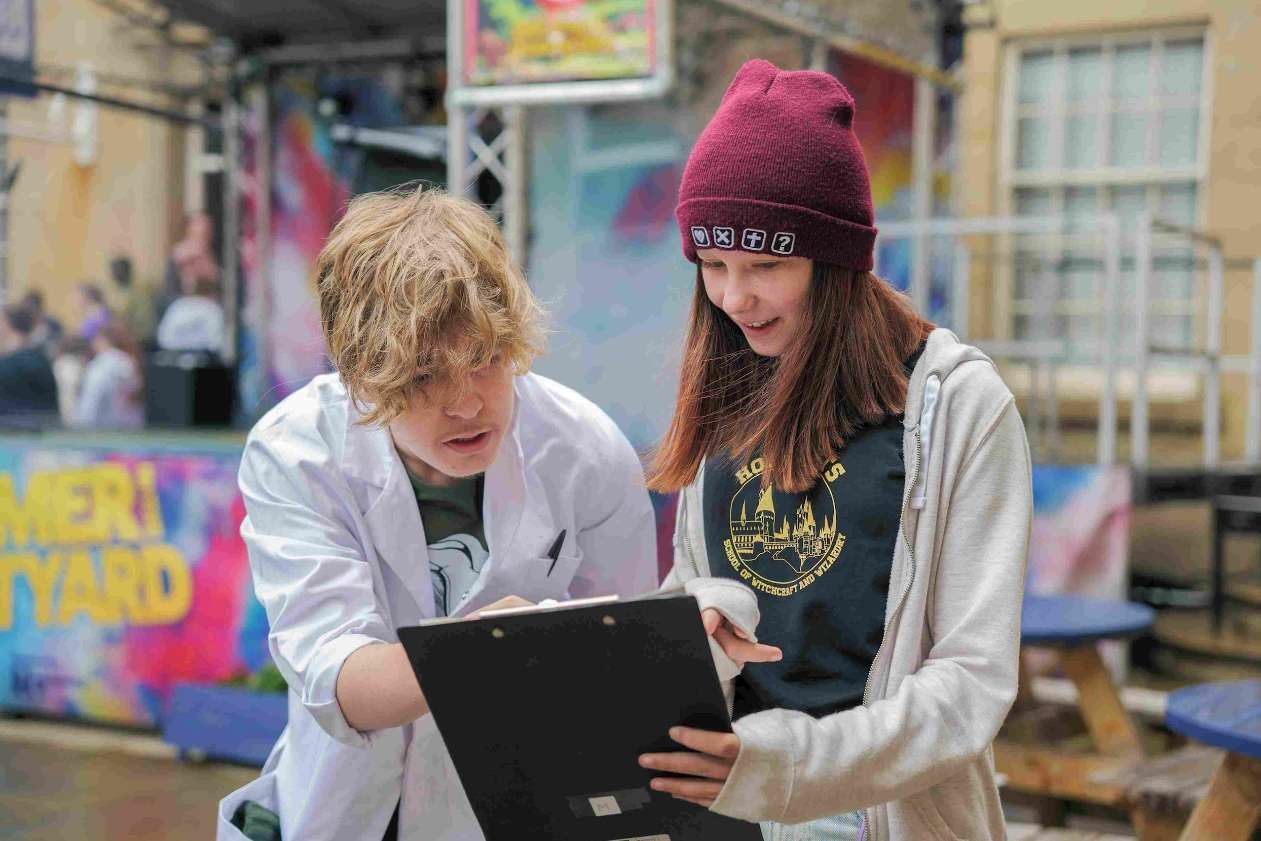 Two teenagers engage with a clipboard in an outdoor setting. One wears a white lab coat and gestures towards the clipboard, while the other, dressed in a hoodie and maroon beanie, smiles while looking at it. A colourful 'SUMMER COURTYARD' sign and blurred background activity suggest a creative or educational event.