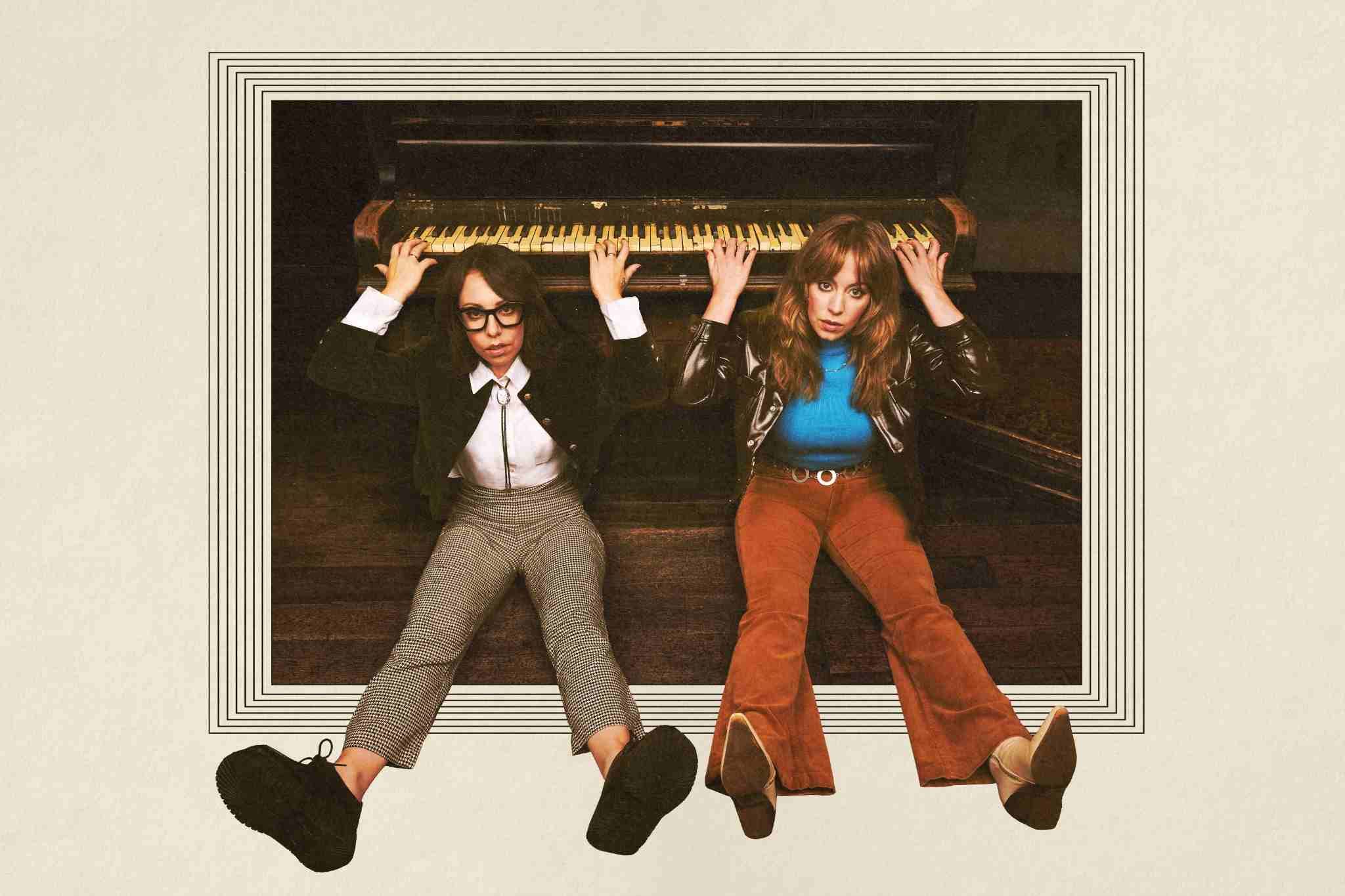 Two people sit on the floor beneath an upright piano, holding onto the piano keys above them. They look directly at the camera with serious expressions, wearing retro-style clothing, framed by a graphic border that gives the image a vintage feel.