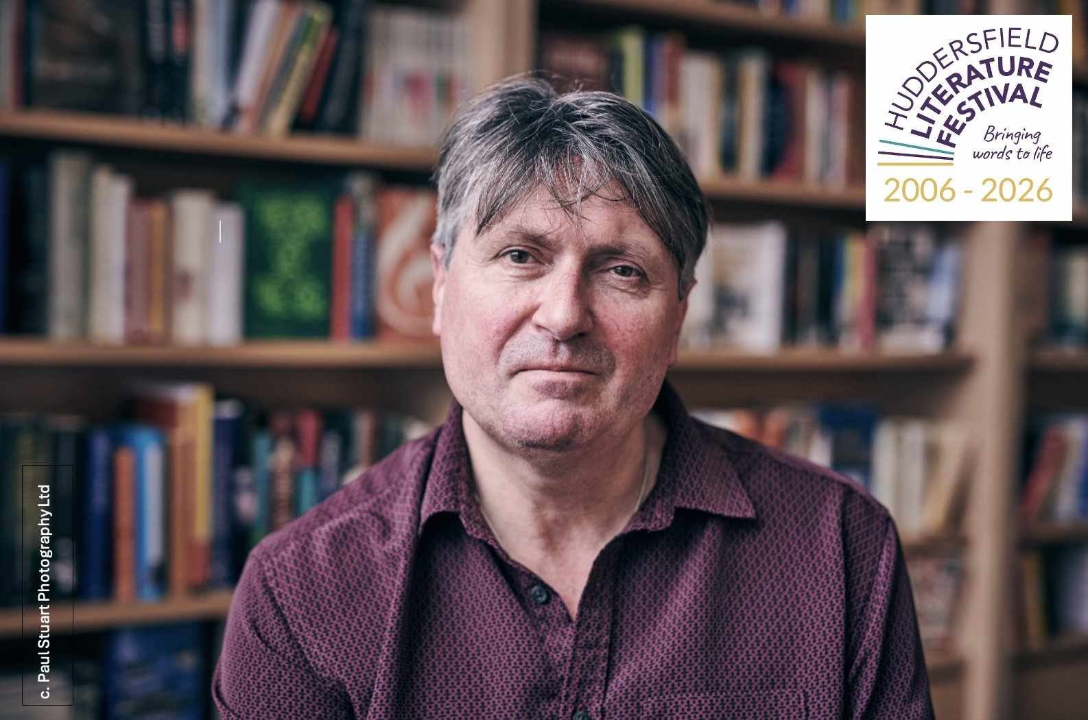 A portrait of a middle-aged man with grey hair wearing a patterned shirt, seated in front of bookshelves filled with books, looking directly at the camera.