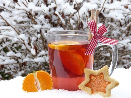 A glass mug of hot fruit punch with orange slices sits on snow, decorated with a red gingham ribbon and clothespin. Beside the mug are a halved clementine and a star-shaped biscuit with a jam centre. Snow-covered branches fill the background.