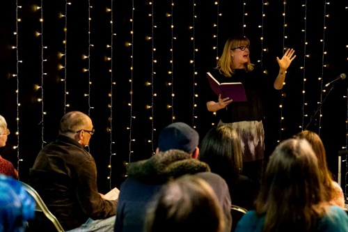 Host, Rose Condo, stands in front of a seated crowd reading from a notebook. 