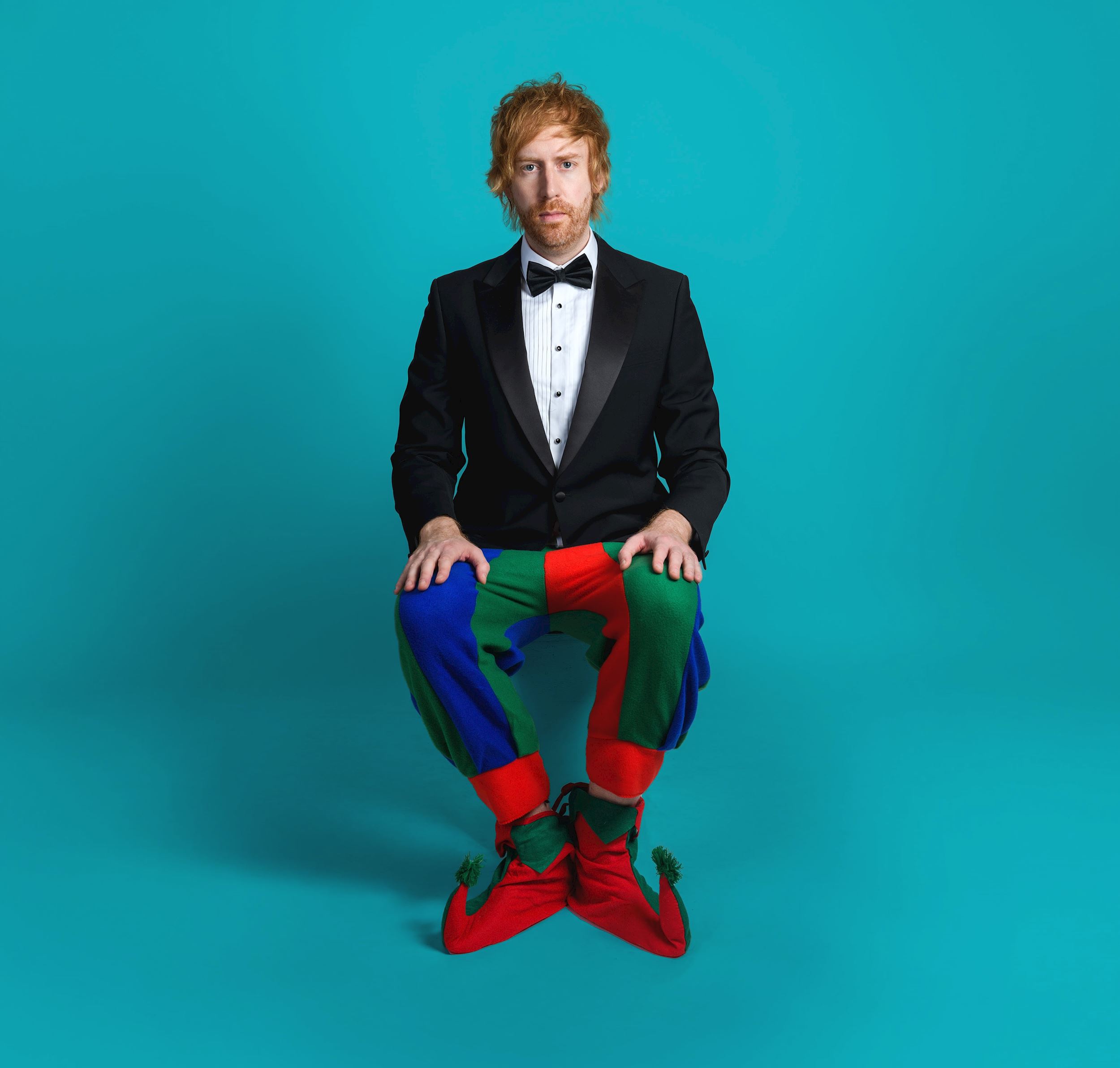 A portrait of comedian Mark Simmons, seated and looking directly at the camera with a neutral expression. He is wearing a formal black tuxedo with a bow tie and a white dress shirt and multi coloured jester trousers, set against a blue background.