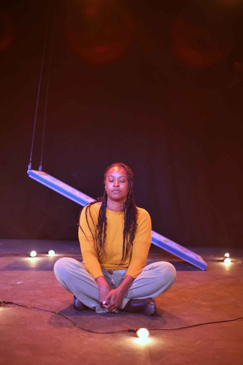 A performer is sat cross-legged on a stage. They are wearing a yellow jumper and grey trousers. 