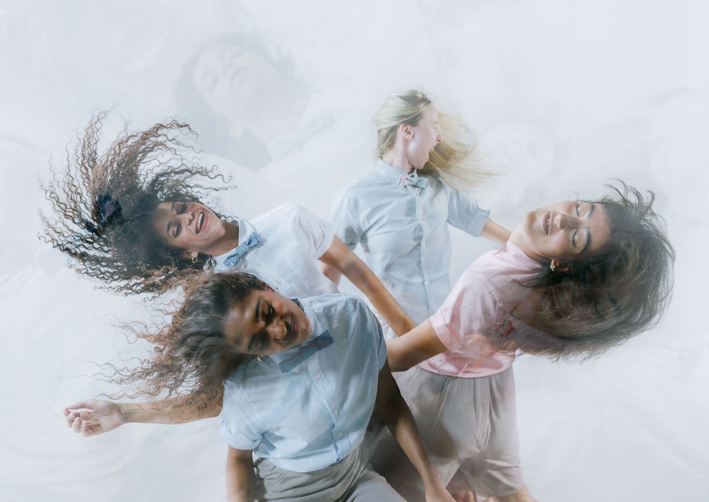 An image of four dancers in pastel shirts and bow ties move expressively with flowing hair and joyful expressions against a soft, misty background.