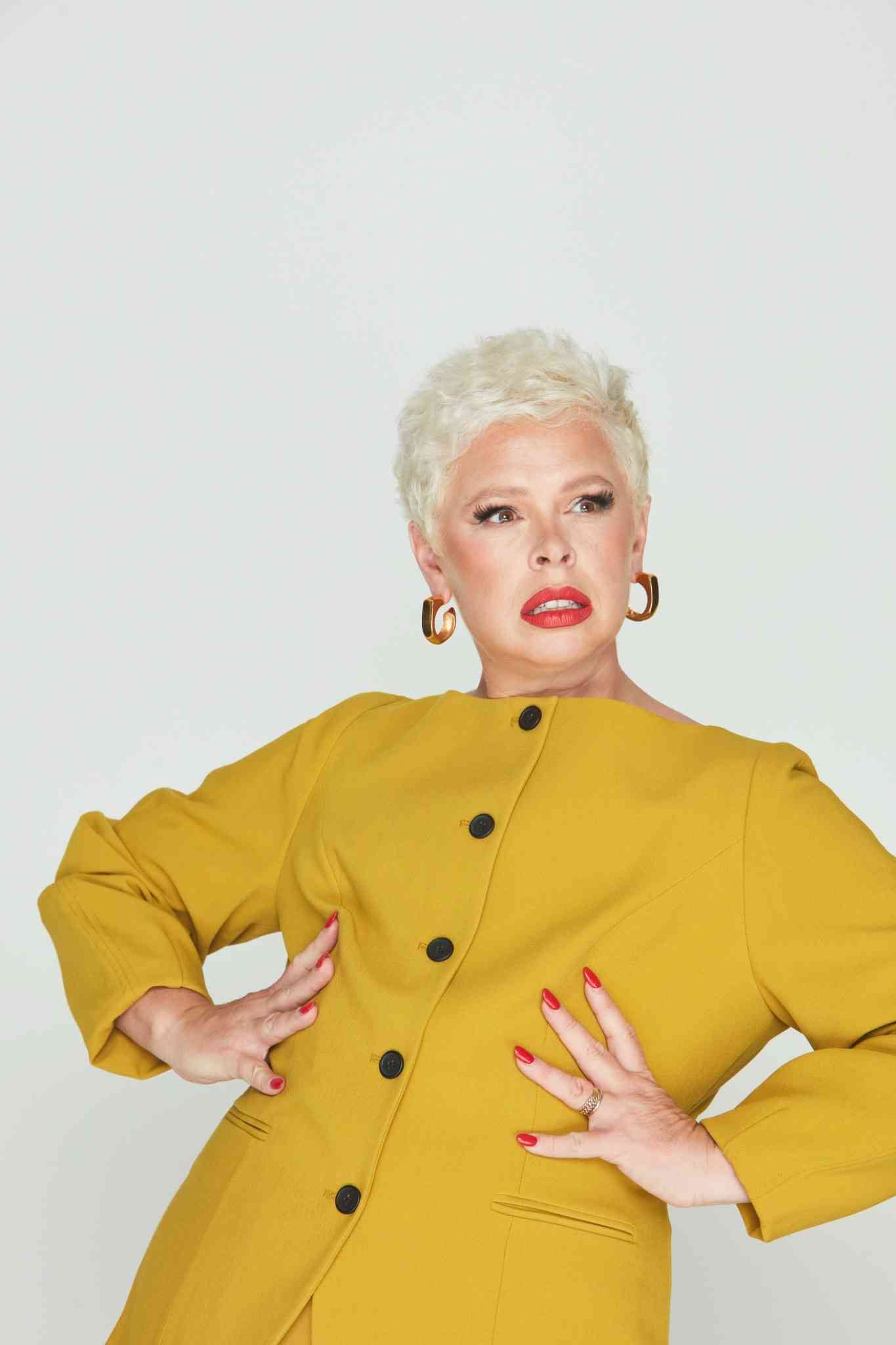 A person with short platinum blonde hair wearing a mustard yellow buttoned jacket, gold hoop earrings, and bold red lipstick, striking a dramatic pose with hands on their waist against a plain light background.