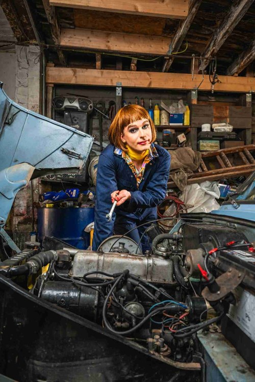 A person in a bob wig and full makeup wearing a mechanic's jumpsuit poses in a cluttered garage, holding a wrench over an open car engine. The scene combines traditional workshop tools with theatrical flair, suggesting a humorous or performative setup.
