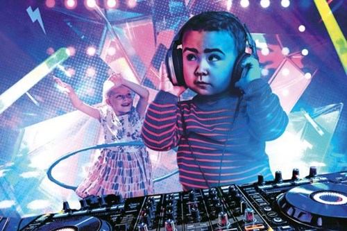 The image features a playful scene of young children in a disco-like environment. In the foreground, a toddler is wearing oversized headphones, standing at a professional DJ deck, as if they are mixing music. The child, dressed in a striped sweater, looks focused and serious while interacting with the equipment. In the background, another child is joyfully dancing, wearing a patterned dress, with arms raised and a hula hoop around their waist.