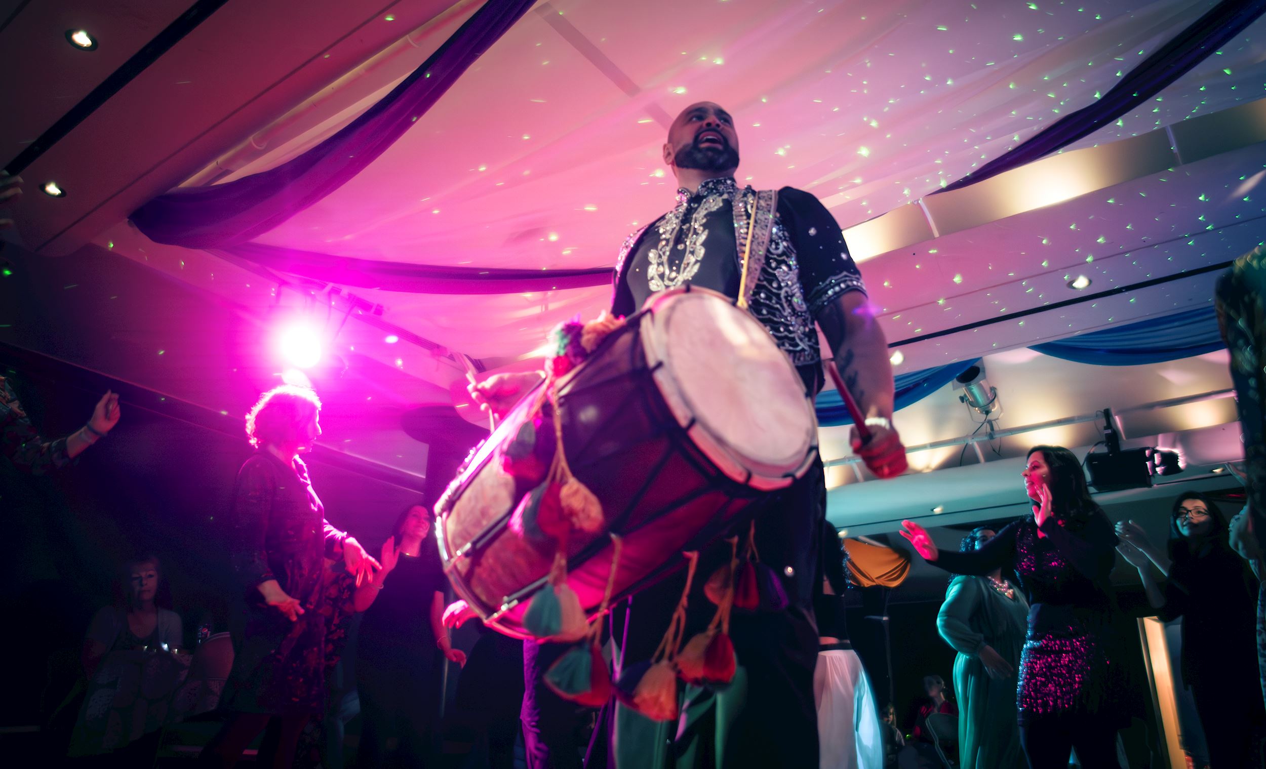 The image captures a vibrant and energetic Bhangra performance in an indoor setting, with colourful lighting creating a festive atmosphere. At the centre, a male performer wearing a traditional, ornate outfit is playing a large dhol drum, with tassels hanging from its sides. His stance is strong and commanding, and the bright stage lights cast a pink and purple glow over him and the room. Surrounding him, people are joyfully dancing, clapping, and moving to the rhythm of the music.