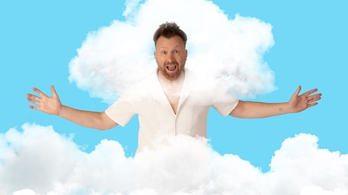 A man with short, styled brown hair and a beard stands against a bright blue background, wearing a cream short-sleeved button-up shirt. He has an excited, open-mouthed expression and extends his arms wide. White fluffy clouds partially obscure his body, blending into his shirt and giving the appearance that he is emerging from or floating within the clouds.