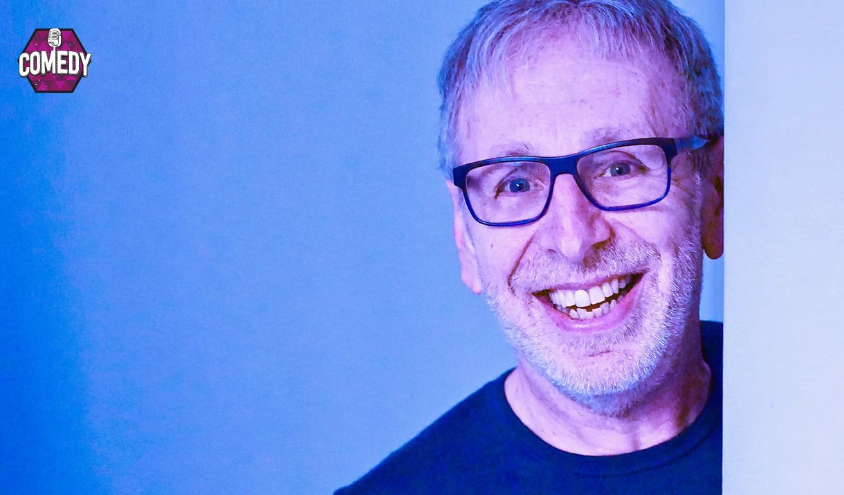 Smiling man with glasses and a grey beard, wearing a dark shirt, peeking playfully from behind a white wall against a blue background. A 'Comedy' badge appears in the top left corner.