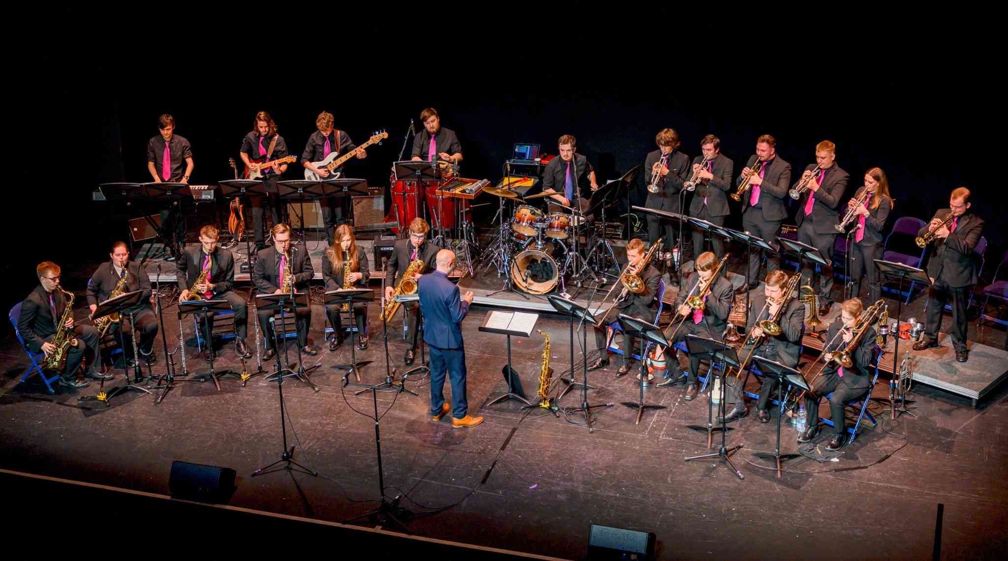 A large jazz band performing on stage, featuring musicians playing brass, woodwind, guitar, bass, drums, and percussion instruments, all dressed in black with pink ties, led by a conductor.