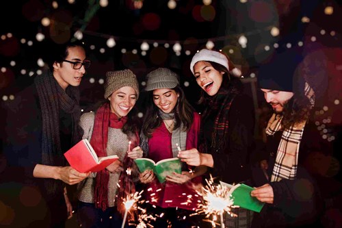 A group of five friends stands outdoors, cheerfully singing Christmas carols together. They are dressed warmly in winter attire, with scarves, hats, and coats. The scene is festive and joyful, illuminated by glowing sparklers they hold in their hands. Each person also holds a songbook.