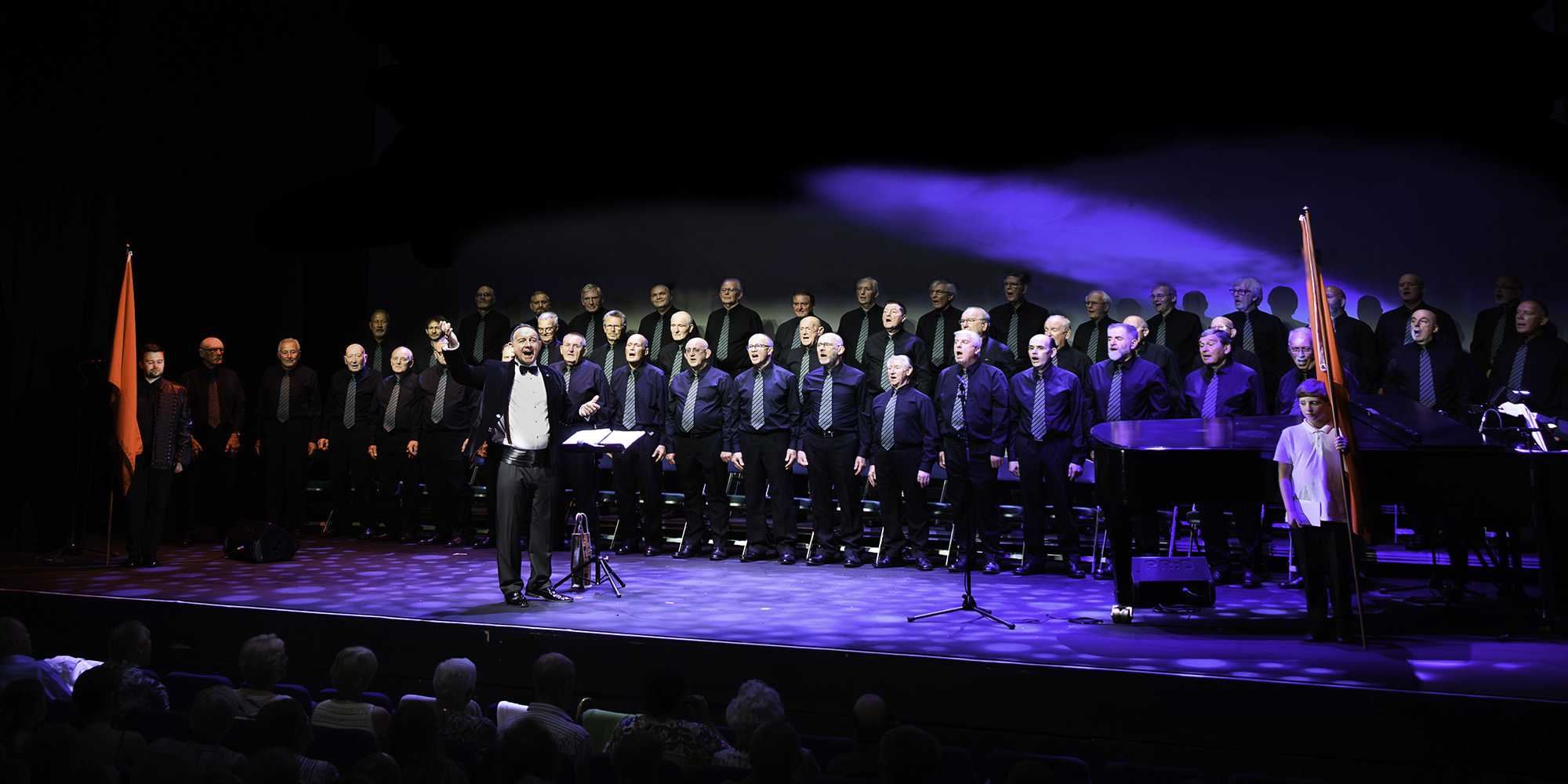 A large male voice choir performs on stage under blue lighting at the Lawrence Batley Theatre. The conductor stands at the front with one arm raised, leading rows of men dressed in black shirts with matching ties. A grand piano sits to one side, and a young flag bearer stands at the front holding a red flag. An audience is visible seated in the foreground.